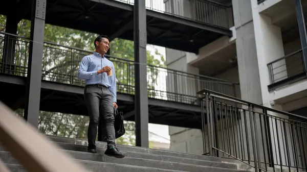 A confident and successful Asian millennial businessman walking down the stairs outside the building with a smile on his face, holding a coffee cup and a briefcase. businesspeople concept