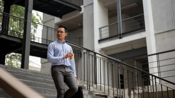 A confident, determined Asian millennial businessman walking down the stairs outside the building with a coffee cup and a briefcase in his hand, looking away from the camera.