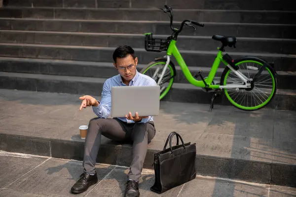 A confused Asian businessman sits on steps with a laptop, showing puzzled expression, perplexed or frustrated, facing a challenging task or problem while working outdoors.
