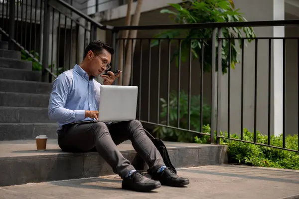 A determined, busy Asian millennial businessman sits on outdoor steps, sending voice message via his smartphone while working on his laptop, working remotely outdoors.