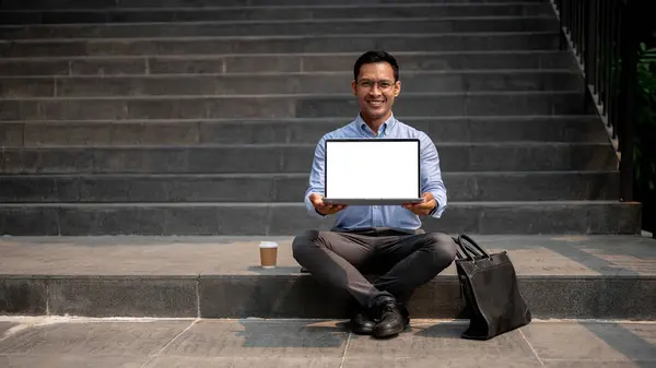 A confident Asian millennial businessman sits on outdoor steps with a coffee cup and a briefcase beside him, holding a laptop with a blank screen. business people concept