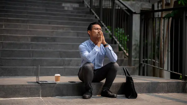 An Asian millennial businessman sits on outdoor steps with a laptop, a briefcase, and a coffee cup beside him, appearing upset and worried, experiencing frustration and the fear of being fired.
