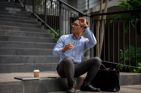 An Asian businessman sits on outdoor steps, looking upward with his hand shielding his eyes from the sun, lost in thought or observing something in the distance.