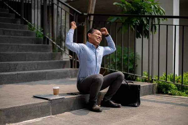 An angry, stressed Asian businessman sits on outdoor steps, crying, screaming, experiencing stress or disappointment, being fired from his job.