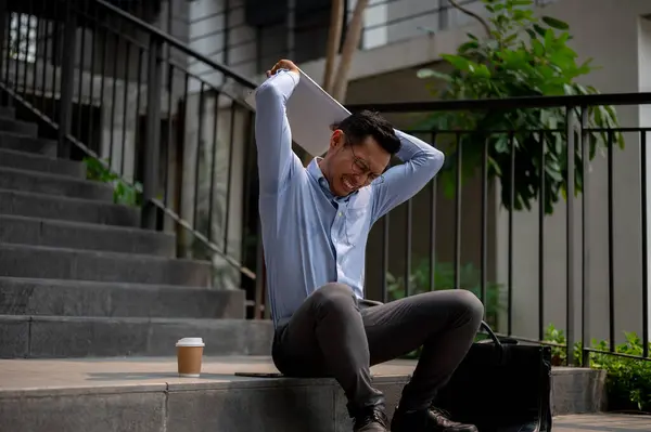 An angry, stressed Asian businessman sits on outdoor steps, holding a laptop with frustration, experiencing stress or disappointment, being fired from his job.