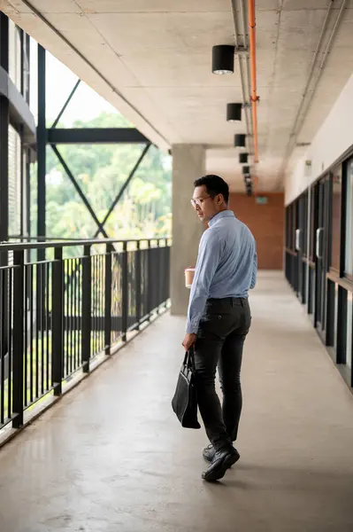 A determined, confident Asian millennial businessman walking along an indoor corridor in the company building, holding a coffee cup and a briefcase. businesspeople concept