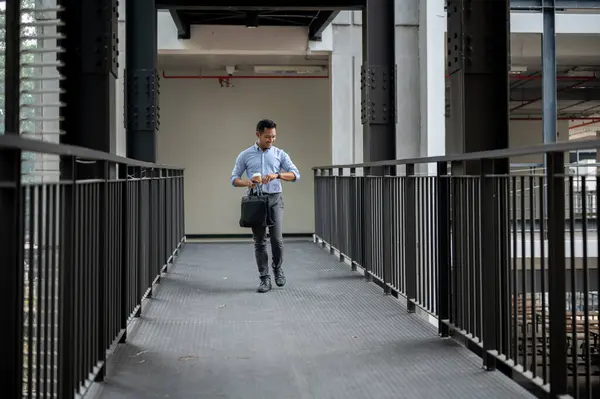 A confident, professional Asian millennial businessman is checking time on his wristwatch while walking along a building corridor, holding a briefcase and a coffee cup. business people concept