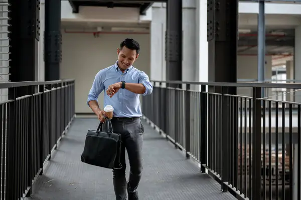 A confident, professional Asian millennial businessman is checking time on his wristwatch while walking along a building corridor, holding a briefcase and a coffee cup. business people concept