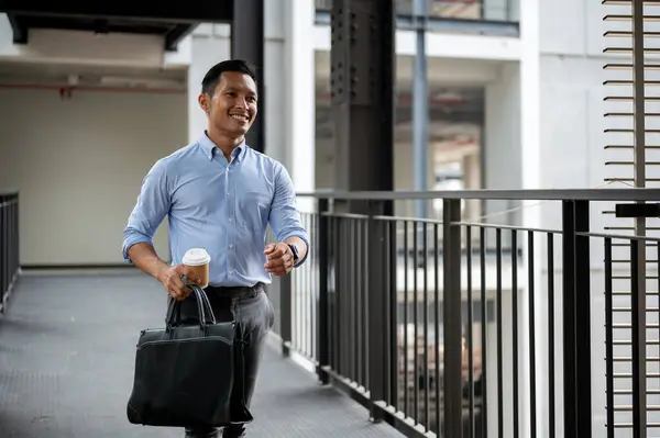 A handsome and confident Asian millennial businessman walking along the building corridor, heading to his office or leave the office, holding a coffee cup and a briefcase.