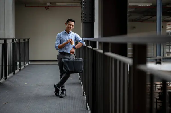 A handsome and confident Asian millennial businessman standing in the building corridor, smiling at the camera, holding a coffee cup and a briefcase, taking a coffee break outdoors.