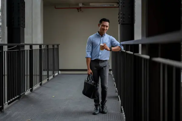 A handsome and confident Asian millennial businessman standing in the building corridor, smiling at the camera, holding a coffee cup and a briefcase, taking a coffee break outdoors.