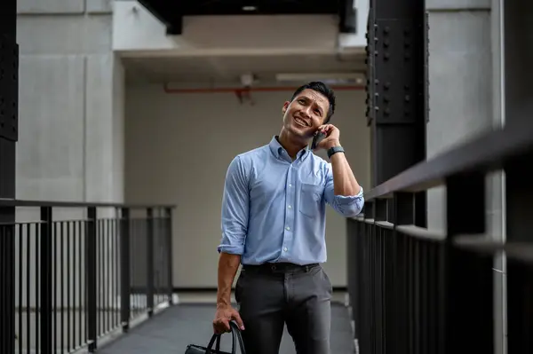 A confused, uncertain Asian millennial businessman talking on the phone with someone while standing on an outdoor corridor in the building. businesspeople and wireless technology concepts
