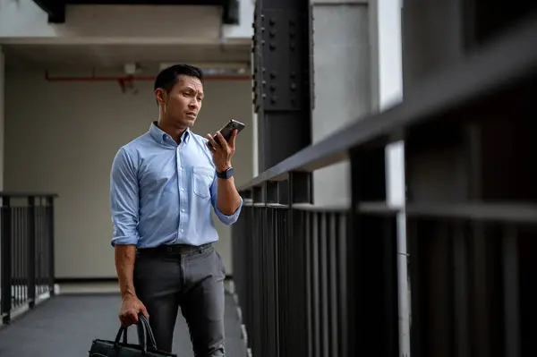 A confused, uncertain Asian millennial businessman checking messages on his smartphone while standing on an outdoor corridor in the building, receiving bad news from his client.