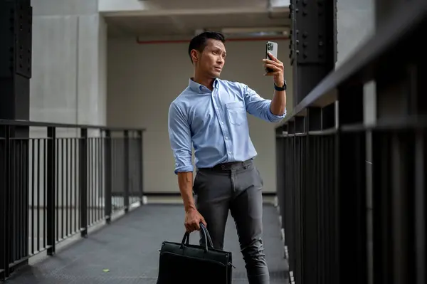 An uncertain Asian millennial businessman looking at his smartphone screen with a confused expression, searching for an internet connection or phone signal while standing on an outdoor corridor.