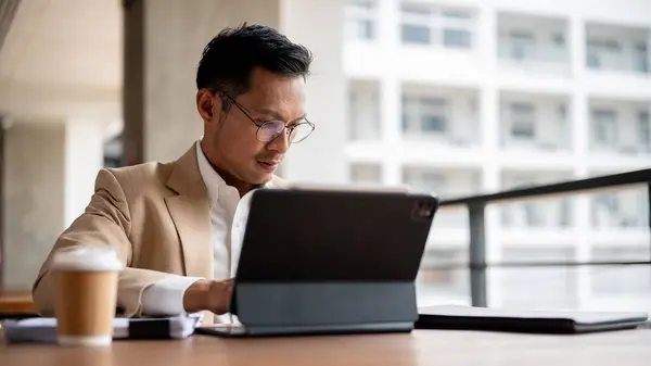 A focused Asian millennial businessman in a beige suit and eyeglasses is working on his digital tablet and reviewing business document at a table in a building corridor.