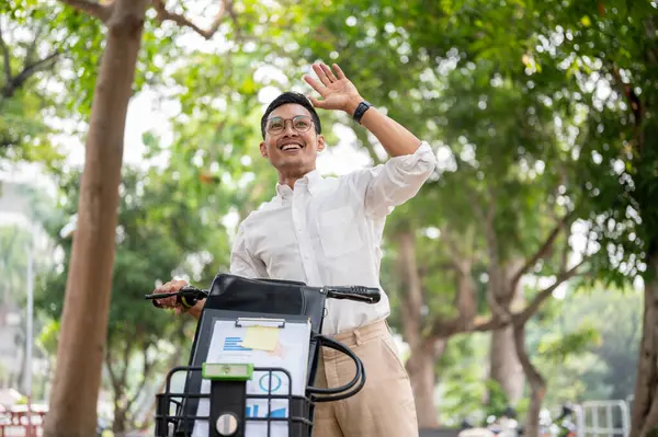 An enthusiastic, happy Asian millennial businessman smiles and waves while pushing his bike in a green park, greeting someone while heading to work in the morning. businesspeople and transportation