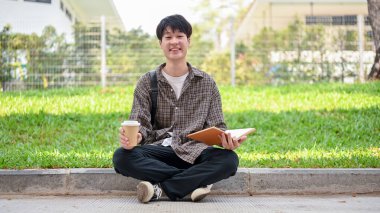Sırt çantası ve günlük kıyafetlerle gülümseyen Asyalı genç bir üniversite öğrencisi elinde kahve fincanı ve kitapla kampüs parkında oturuyor..