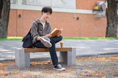 Mutlu, rahat, Asyalı genç bir üniversite öğrencisi kampüs parkında bir bankta oturmuş ders kitabı okuyor ve açık havada eğleniyor. Üniversite hayatı, hobi, yaşam tarzı
