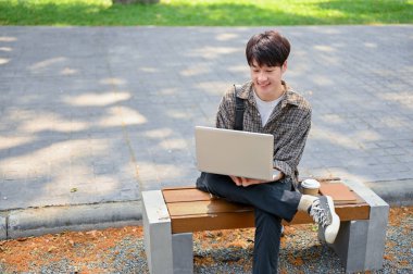 Genç bir Asyalı üniversite öğrencisi kampüs parkındaki bankta dizüstü bilgisayarını kullanıyor. Üniversite hayatı ve kablosuz teknoloji kavramları