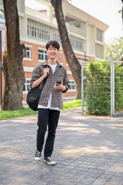 Sırt çantası ve günlük giysiler içinde gülümseyen mutlu bir Asyalı erkek üniversite öğrencisi kampüs parkında yürürken akıllı telefonunu kullanıyor. Üniversite hayatı, yaşam tarzı, kablosuz teknoloji