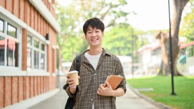 Sırt çantası taşıyan, gülümseyen, pozitif Asyalı genç bir üniversite öğrencisi elinde kahve fincanı ve kitapla kampüste tuğla bir binanın önünde duruyor..
