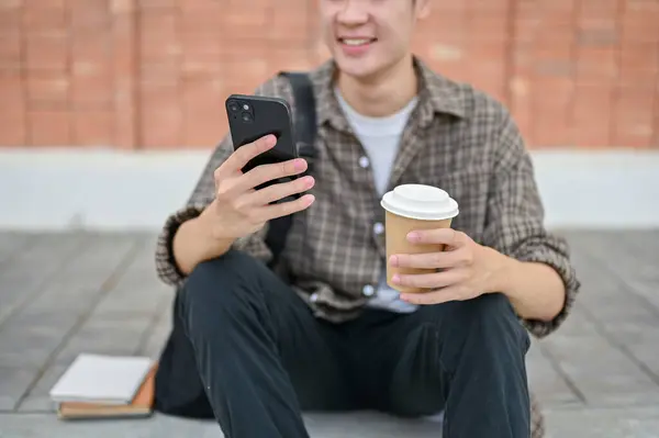 A cropped image of a young male college student sits on the street near the campus building using his smartphone and holding a coffee cup. wireless technology and lifestyle concepts