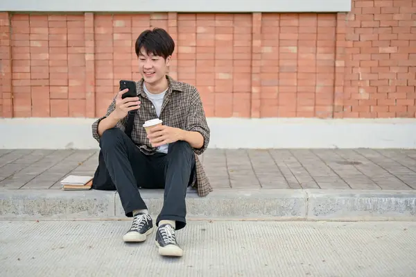 A handsome, happy young Asian male college student sits on the street in a campus, using his smartphone and holding a coffee cup, chatting with his friends or scrolling on his social media.