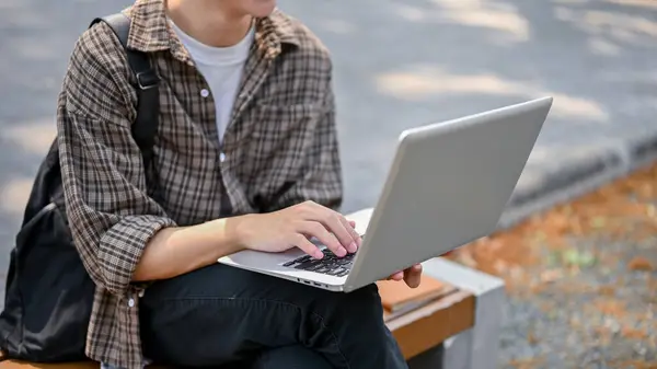 A cropped image of a male college student in casual wear using his laptop computer on a bench in the campus park. university life and wireless technology concepts