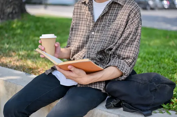 A cropped image of a male college student in casual wear sits on a stone bench in a park, holding an open book and a cup of coffee, reading a book outdoors.