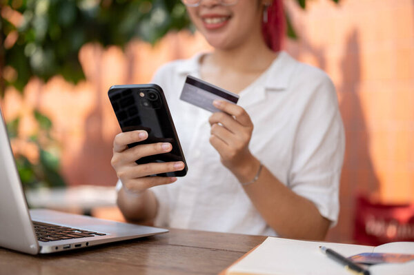 A cropped image of an Asian woman holding a smartphone and a credit card while sitting at a table outdoor, making an online payment, shopping online, transferring money on a mobile app