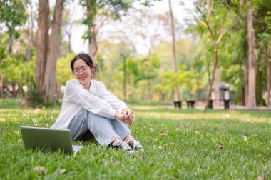 Gözlüklü mutlu bir Asyalı kadın parkta çimenlerde oturur, bilgisayarına bakarken gülümser, aydınlık bir günde parkta uzaktan çalışır. kablosuz teknoloji ve yaşam tarzı kavramları