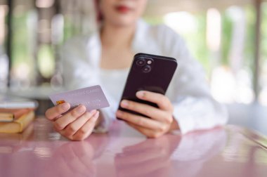 Sıradan giyinmiş Asyalı bir üniversite öğrencisi kafeteryadaki bir masada elinde kredi kartı ve akıllı telefonuyla gezici bir bankacılık uygulaması ile oturuyor. çevrimiçi ödeme, online alışveriş