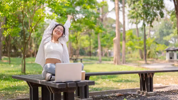 A young carefree, relaxed Asian woman in casual wear sits on a bench in a green park on a bright day, leans back, stretching her arms, enjoying nature while working remotely.