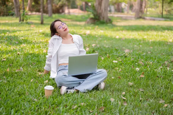 A happy, relaxed Asian woman in glasses sits on the grass in a park with her laptop, enjoying the sunlight, leaning back with her eyes closed. lifestyle and wireless technology concepts