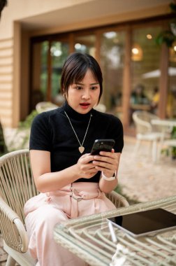 Şok olmuş Asyalı bir kadın akıllı telefon ekranına bakıyor çarpıcı bir yüzle, telefonunda bir mesajla, açık bir kafede oturuyor..