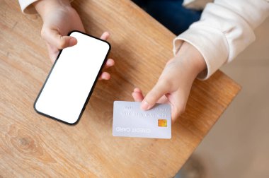 Kredi kartı ve akıllı telefonu ahşap bir masanın üzerinde tutan bir kadının en iyi görüntü görüntüsü. Grafik reklamlarınızı göstermek için akıllı telefon beyaz ekran modeli. Çevrimiçi alışveriş, çevrimiçi ödeme, nakit yok