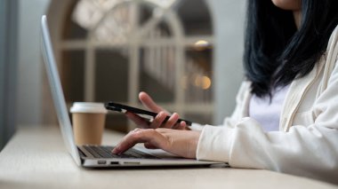 Akıllı telefon ve dizüstü bilgisayar kullanan, dizüstü bilgisayarda klavyeye yazan, kafede uzaktan çalışan Asyalı bir kadının kırpılmış görüntüsü. İnsanlar ve kablosuz teknoloji kavramları