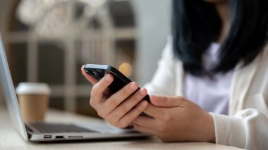 Ofis masasında ya da laptopuyla masada akıllı telefonunu kullanan Asyalı bir kadının yakın plan görüntüsü. Mesajları kontrol etmek, sohbete cevap vermek, metin okumak, insanlar ve teknoloji kavramları