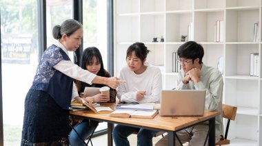 Yetişkin bir Asyalı bayan öğretmen sınıftaki öğrencilerine ders veriyor, son proje detaylarını anlatıyor, öğrencilerine yardım ediyor ve soruları cevaplıyor..