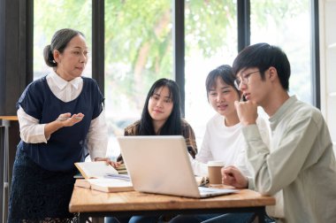 Yetişkin bir Asyalı bayan öğretmen sınıftaki öğrencilerine ders veriyor, son proje detaylarını anlatıyor, öğrencilerine yardım ediyor ve soruları cevaplıyor..