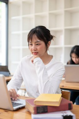Güzel bir Asyalı genç bayan öğrenci dizüstü bilgisayarıyla işine odaklanıyor, kampüsteki kütüphanede ortak çalışma alanında projesi üzerinde çalışıyor. Eğitim ve yaşam tarzı kavramları