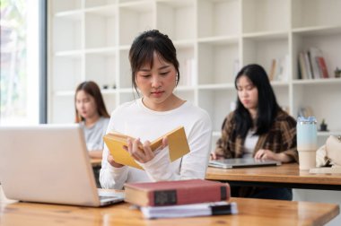 Genç ve güzel Asyalı bir kız öğrenci dizüstü bilgisayarıyla işine odaklanıyor, bir kitaptaki bilgileri araştırıyor, kampüste bir kütüphanede ortak çalışma alanında projesi üzerinde çalışıyor..