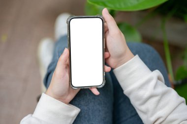 Günlük giysiler içinde bir ev bitkisinin yanında oturan ve akıllı telefonunu kullanan bir kadının yakın plan görüntüsü. Beyaz ekran akıllı telefon maketi. sohbet, sosyal medya, online alışveriş, mobil uygulama