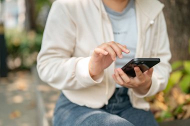 Sıradan giyinmiş genç bir Asyalı kadının şehir parkındaki bir bankta oturup akıllı telefonunu kullanırken çekilmiş bir fotoğrafı. sohbet etmek, mobil uygulama kullanmak, mesajları kontrol etmek, sosyal medyada kaydırmak, online alışveriş