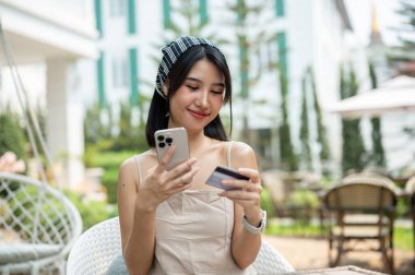 Sevimli, gülümseyen, minimal elbiseli Asyalı bir kadın güzel bir İngiliz bahçesinde elinde kredi kartı ve akıllı telefonuyla bir masada oturmuş internetten alışveriş yapmaktan zevk alıyor. mobil bankacılık, çevrimiçi ödeme