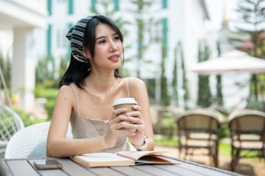 Şirin bir elbise giymiş çekici bir Asyalı kadın elinde kahve fincanı ile kameradan uzağa bakıyor ve güzel bir bahçede bir masada otururken bir şeyler düşünüyor ya da hayal kuruyor..