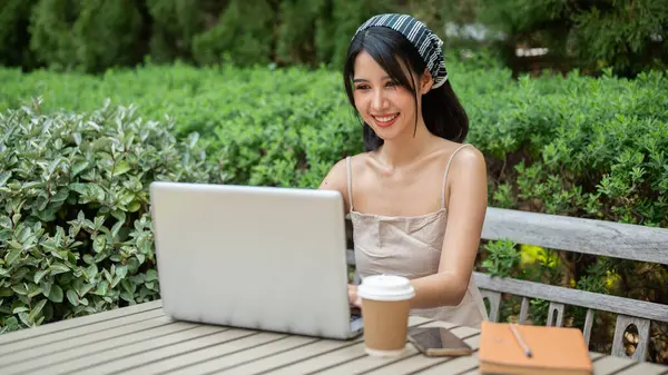 An attractive Asian woman in a cute minimal dress is working on her laptop computer at a table in a green garden backyard, working remotely outdoors. people and technology concepts