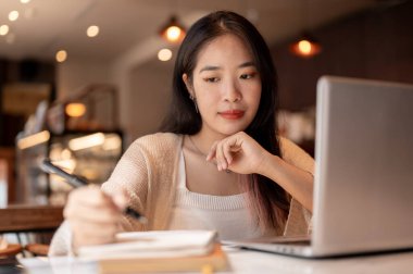 Genç odaklı Asyalı kadın bir kafede uzaktan çalışıyor dizüstü bilgisayarıyla masada otururken defterine notlar alıyor. İnternette okuyan ve bir kafede ödev yapan bir öğrenci..