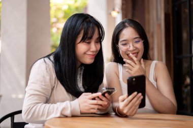 İki mutlu genç Asyalı kadın, şehirdeki bir kafe veya restoranın açık masasında otururken akıllı telefondan konuşmaktan ve bir şeyler izlemekten zevk alıyorlar. insanlar ve yaşam tarzı kavramları