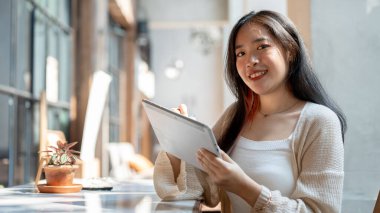 Gündelik giyinmiş güzel bir Asyalı kadın dijital tablet tutuyor ve güneşli bir günde açık bir kafede otururken kameraya gülümsüyor..